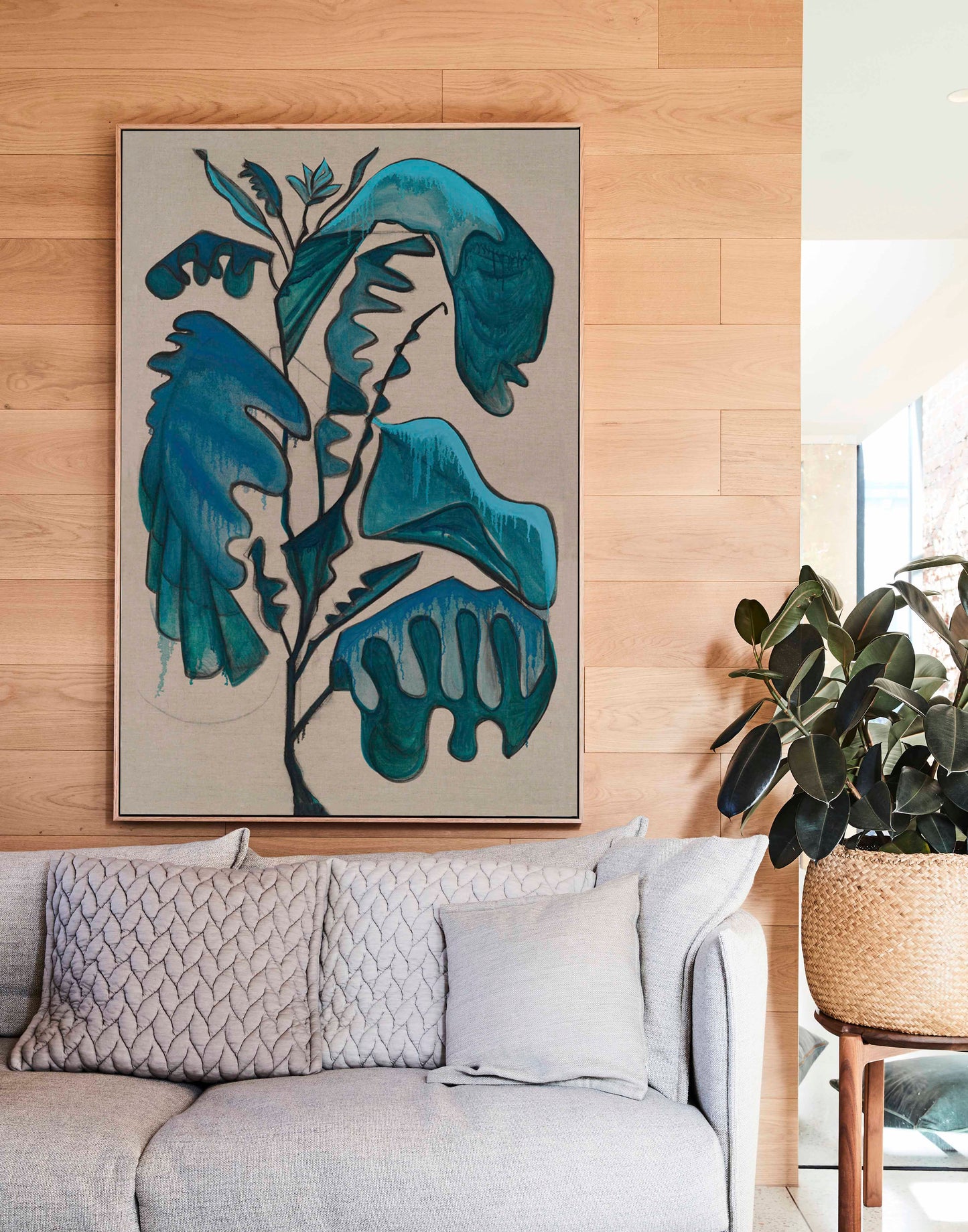 Studio Elwood “Terracotta Leaf” — large framed botanical artwork in blue-green tones displayed above a modern neutral sofa with warm timber finishes and indoor greenery.