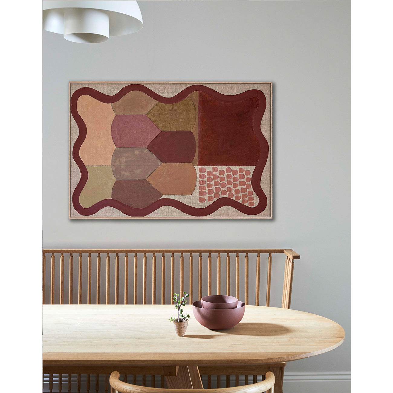 Abstract framed artwork in warm earth tones hanging above a minimalist wooden dining table with a spindle-back bench, small plant, and ceramic bowls beneath soft pendant lighting.